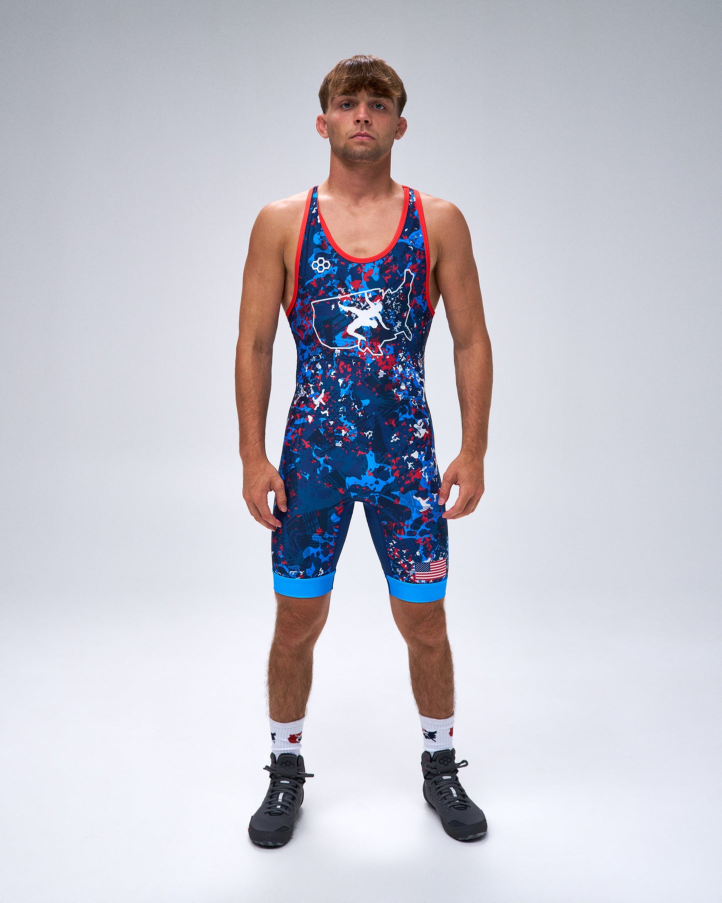 A male model stands against a light gray studio background wearing a sleeveless athletic wrestling singlet with a blue, red, and white abstract pattern, red trim, white socks, and black cleats.