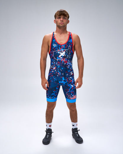 A male model stands against a light gray studio background wearing a sleeveless athletic wrestling singlet with a blue, red, and white abstract pattern, red trim, white socks, and black cleats.