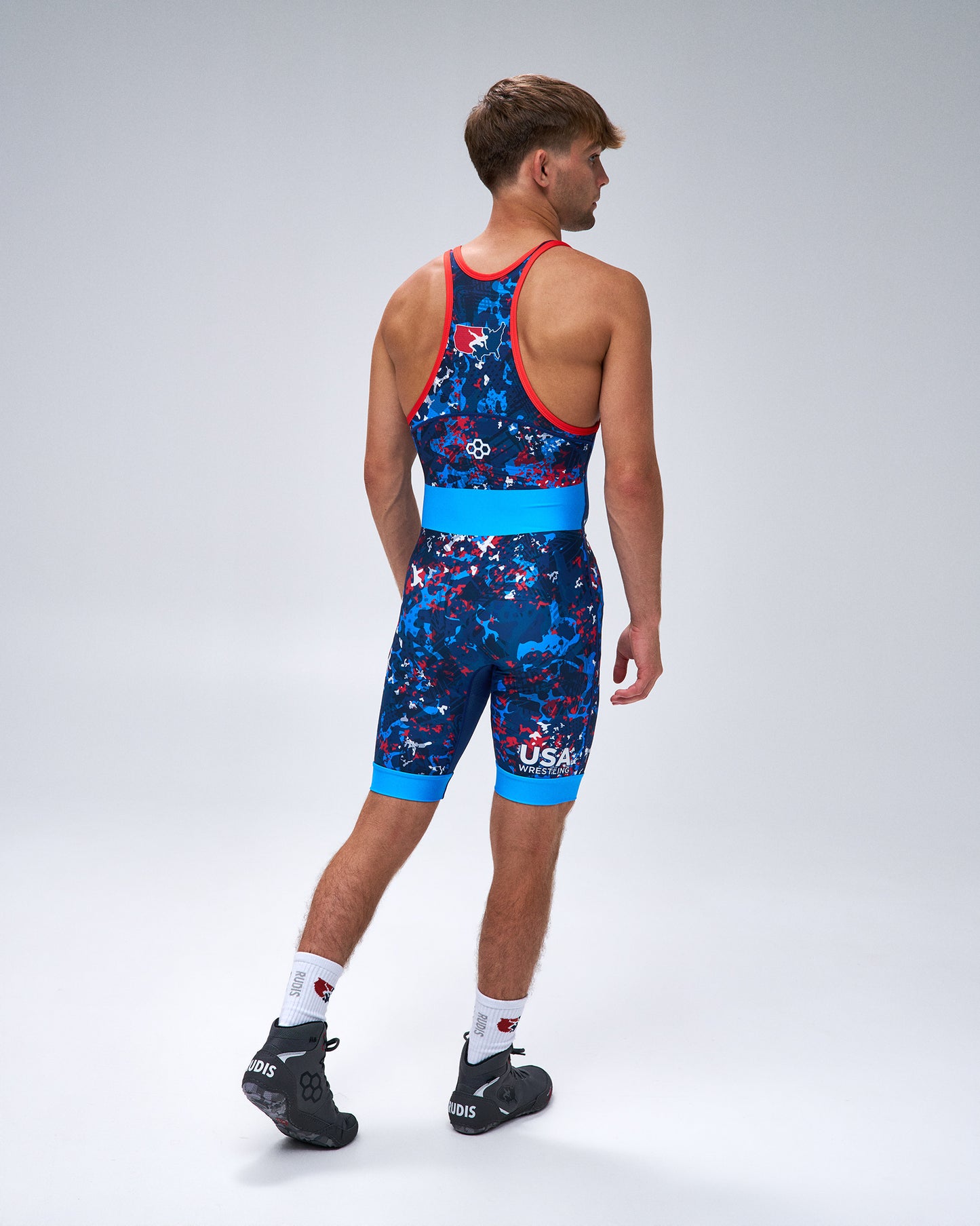 A male model shown from the back wearing a USA Wrestling Singlet with a blue, red, and white abstract pattern, bright blue waistband, and red shoulder straps, paired with white socks and black cycling shoes, on a light gray studio background.