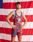 A man modeling a red, blue, and orange patriotic wrestling singlet in front of a large American flag backdrop.