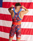 A man shown from behind wearing a patriotic red, white, and blue wrestling singlet with an American flag backdrop.