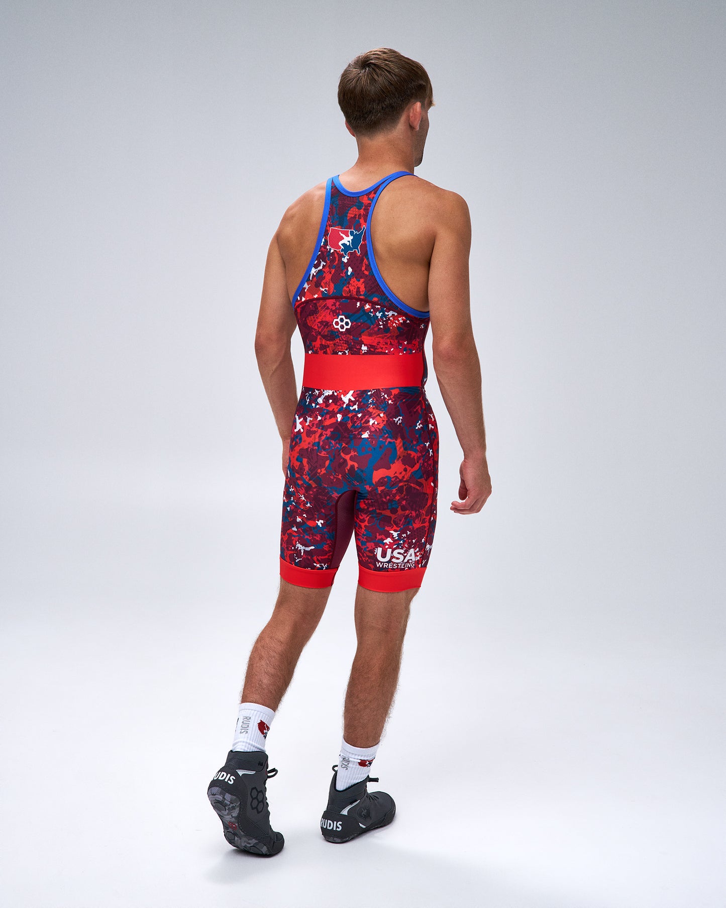 A man shown from the back wearing a red and navy USA Wrestling Singlet with a blue waistband and shoulder straps, paired with white socks and black shoes, against a light gray studio background.