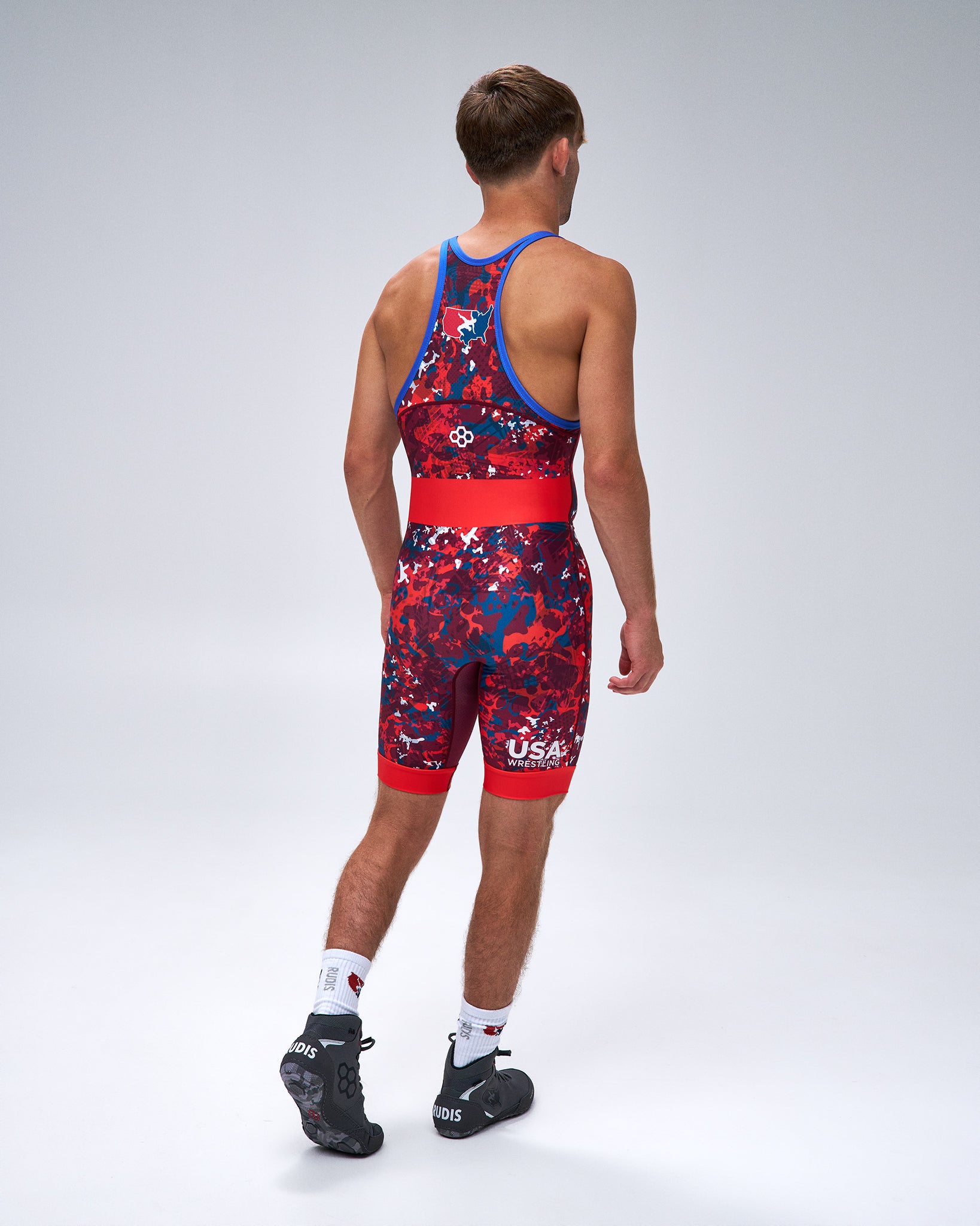 A man shown from the back wearing a red and navy USA Wrestling Singlet with a blue waistband and shoulder straps, paired with white socks and black shoes, against a light gray studio background.