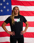 Female athlete wearing the RUDIS USA Wrestling Pastel Logo Elite Super Soft T-Shirt stands confidently with hands on hips in front of an American flag, paired with black athletic leggings.