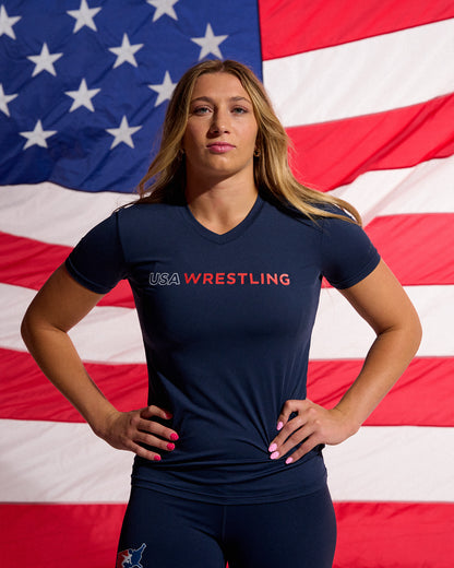 Female athlete wearing the RUDIS USA Wrestling Women's Navy Performance V-Neck T-Shirt stands confidently in front of an American flag with matching USA Wrestling leggings and RUDIS training shoes.