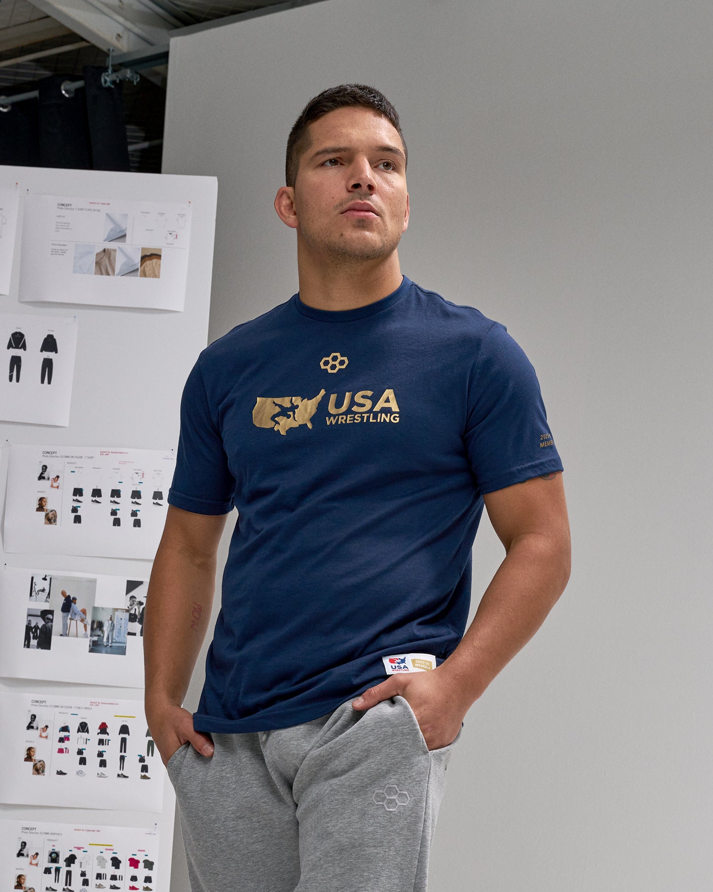 Man wearing a blue 'USA Wrestling' t-shirt standing in front of a wall with design boards.