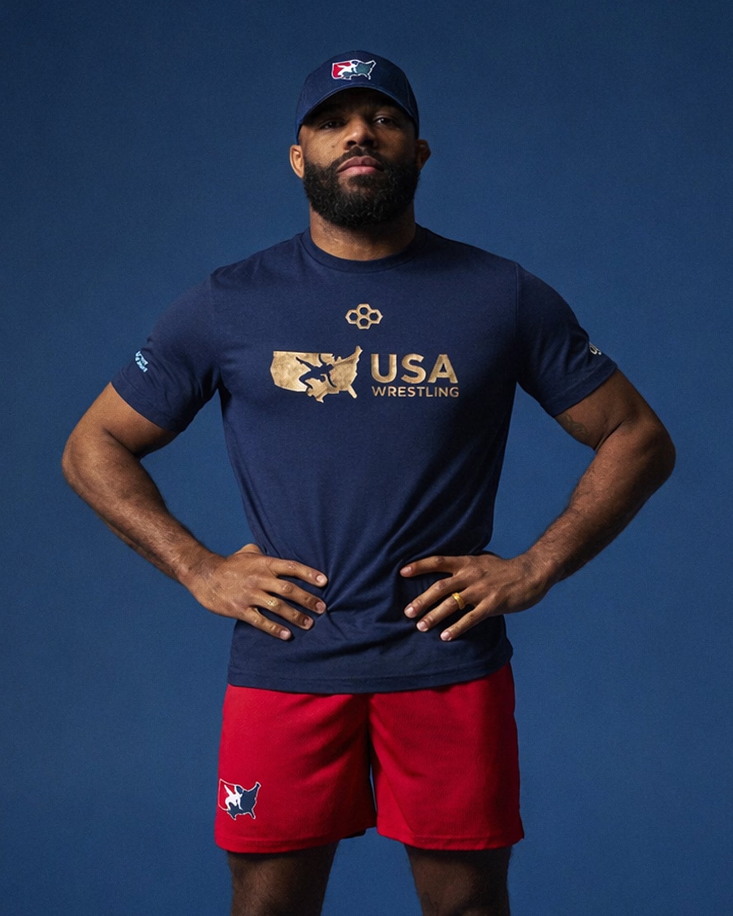 Olympic wrestler Jordan Burroughs wearing a navy 'USA Wrestling' t-shirt and red RUDIS shorts on a blue background