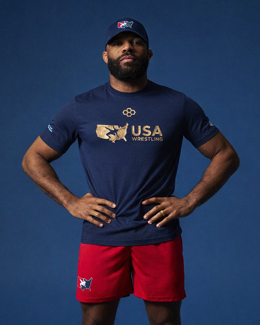 Olympic wrestler Jordan Burroughs wearing a navy 'USA Wrestling' t-shirt and red RUDIS shorts on a blue background