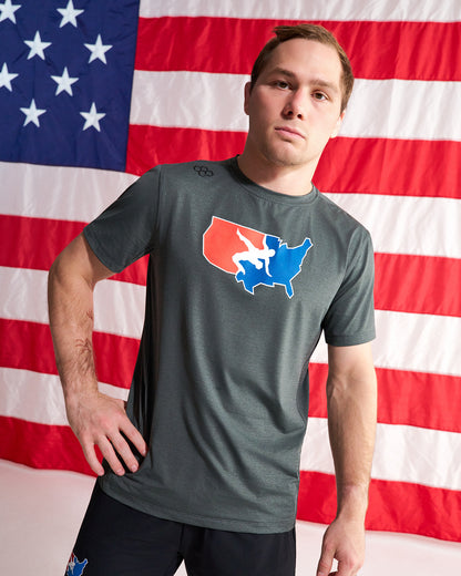 A man wearing a dark gray athletic t-shirt with a red, white, and blue USA map graphic on the chest, posed in front of a large American flag backdrop.