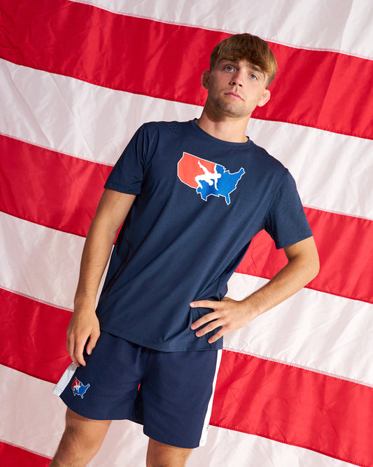 A man wearing a navy blue t-shirt and matching shorts with patriotic red, white, and blue graphics, posed in front of a large American flag backdrop.