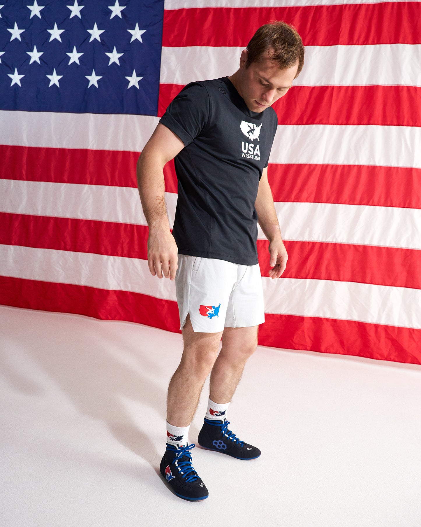 A man wearing a black USA boxing team t-shirt, white boxing shorts, and blue boxing shoes stands in front of a large American flag backdrop.