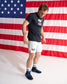 A man wearing a black USA boxing team t-shirt, white boxing shorts, and blue boxing shoes stands in front of a large American flag backdrop.