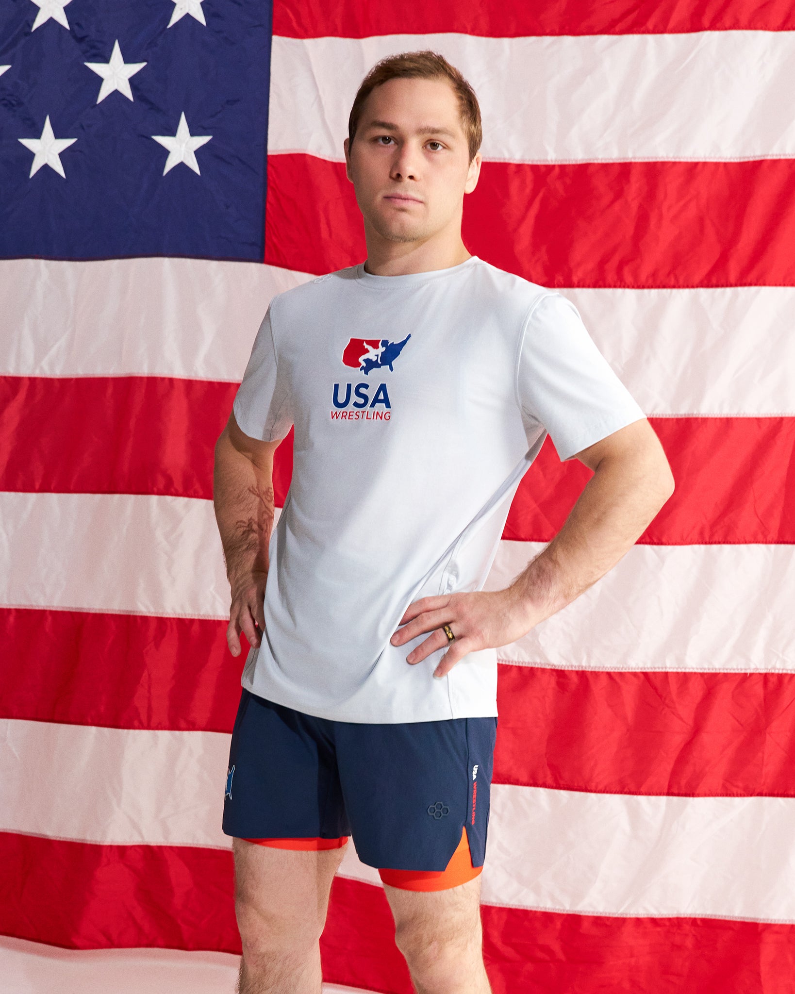A man stands in front of a large American flag backdrop wearing a white USA Wrestling t-shirt and navy athletic shorts with orange lining.
