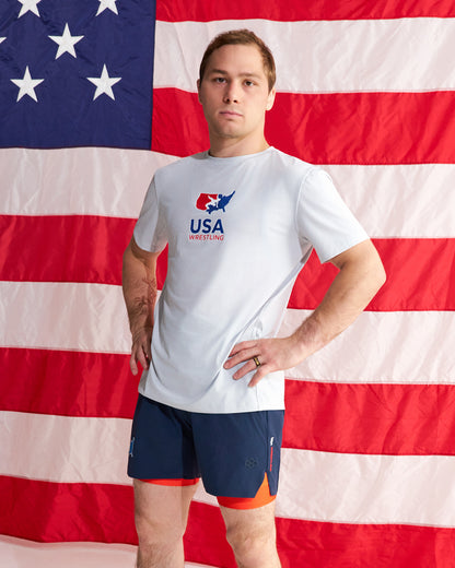 A man stands in front of a large American flag backdrop wearing a white USA Wrestling t-shirt and navy athletic shorts with orange lining.