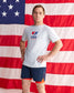 A man stands in front of a large American flag backdrop wearing a white USA Wrestling t-shirt and navy athletic shorts with orange lining.