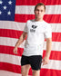 A man wearing a white USA Wrestling t-shirt and black athletic shorts stands in front of a large American flag backdrop.