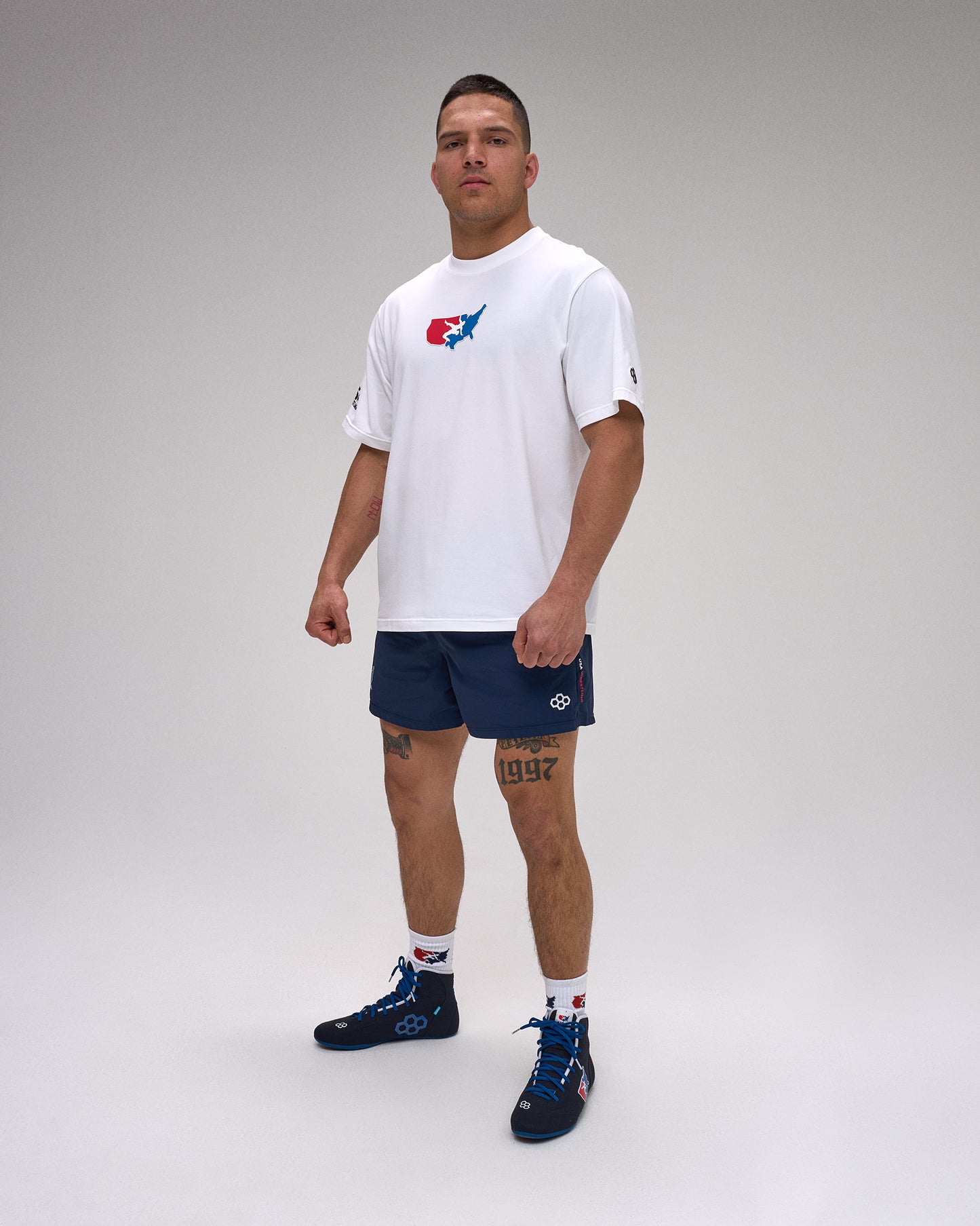 Man wearing a white t-shirt with a 'USA Wrestling' logo and navy shorts on a plain background