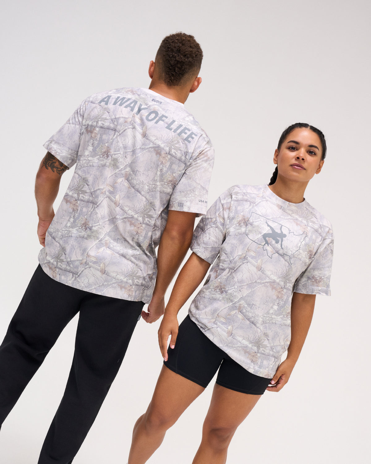 Male and female models wearing the USA Wrestling Map Classic Cotton T-Shirt in tree camo, featuring a gray wrestling graphic over a USA map outline on the front and A WAY OF LIFE lettering across the back.