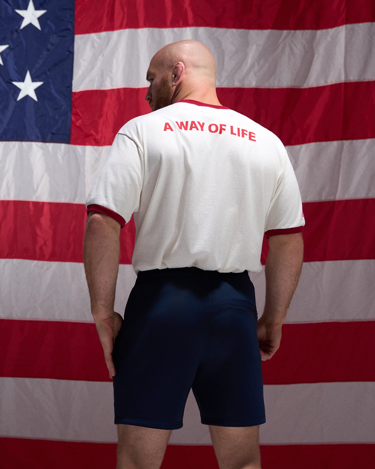 Man wearing a white shirt with 'A Way of Life' text in red, standing in front of an American flag.