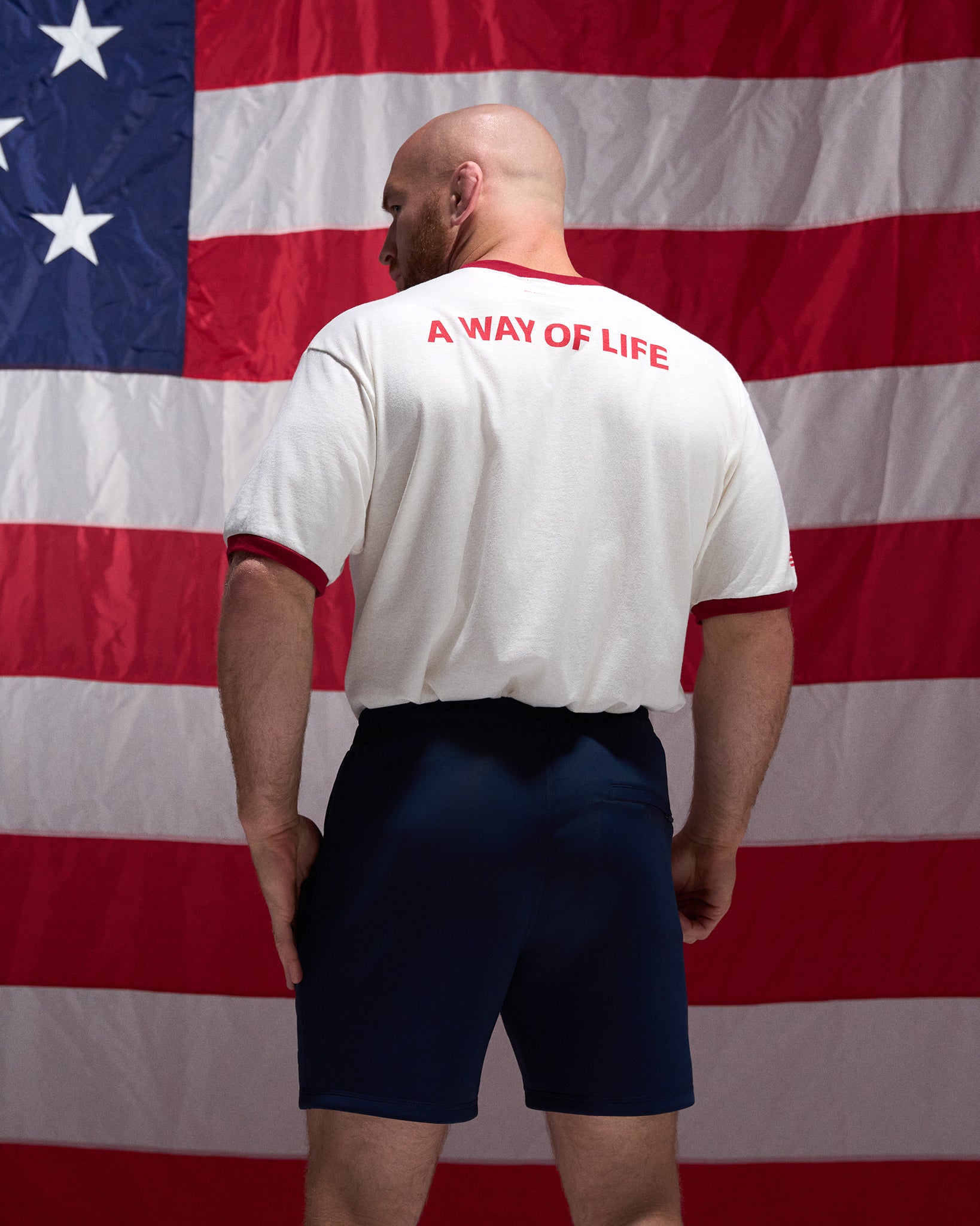Man wearing a white shirt with 'A Way of Life' text in red, standing in front of an American flag.