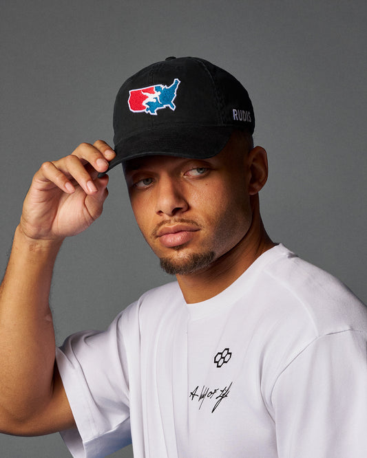 Man wearing a black cap with a logo and text, holding it with his right hand against a gray background.