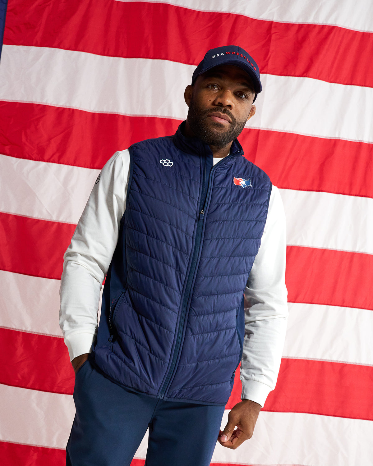 A man wearing a navy blue quilted zip-up vest over a white long-sleeve shirt and navy pants, standing in front of an American flag. The vest has a small white logo on the left chest and a small red, white, and blue logo on the right chest.