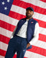 A man wearing a navy blue quilted vest, matching navy pants, a white long-sleeve shirt, and a navy cap stands in front of a large American flag backdrop.