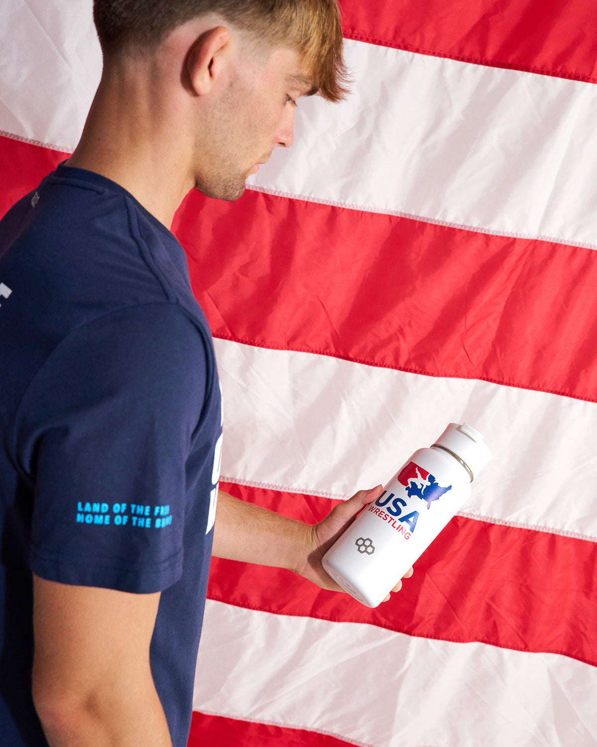 A man in a navy blue t-shirt holding a white water bottle with USA Wrestling branding, set against a large American flag background.