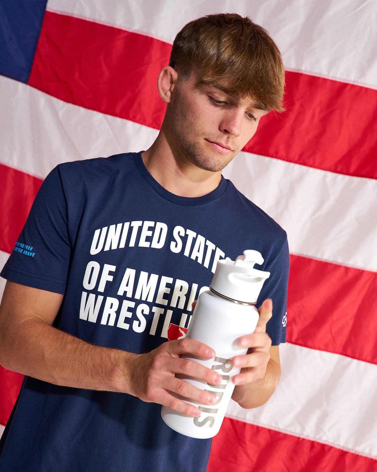 A man wearing a navy blue t-shirt with 'UNITED STATES OF AMERICA WRESTLING' printed in white, holding a white water bottle in front of an American flag background.