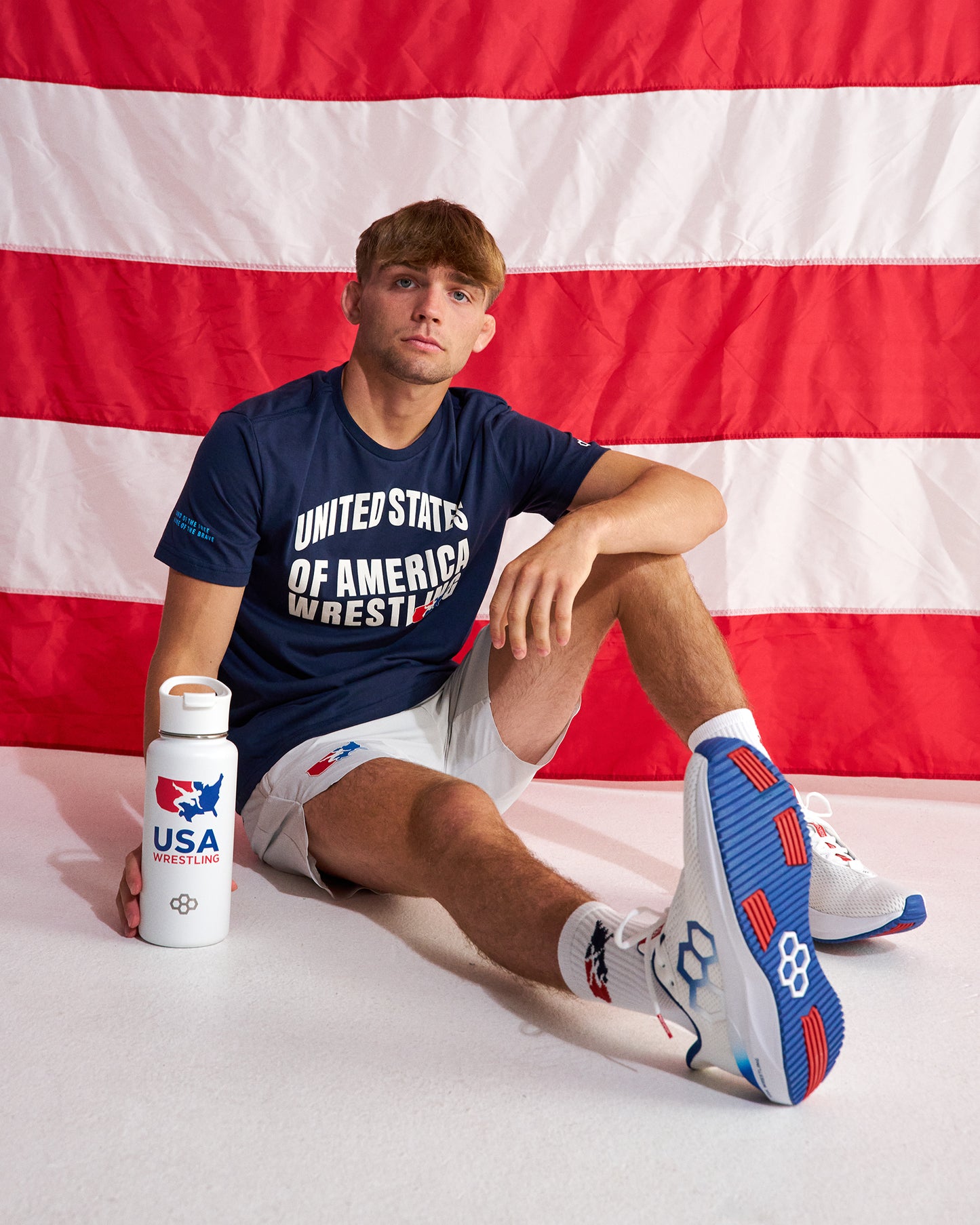 A man sits on a white floor in front of a large American flag backdrop, wearing a navy blue t-shirt with 'UNITED STATES OF AMERICA WRESTLING' printed on the front, white shorts, and white sneakers with blue and red accents. He is holding a white water bottle with a USA Wrestling logo.