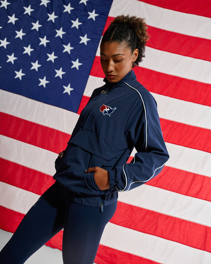 A woman modeling a navy blue athletic tracksuit with white piping and a small red, white, and blue logo on the chest, posed in front of a large American flag backdrop.