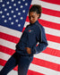 A woman modeling a navy blue athletic tracksuit with white piping and a small red, white, and blue logo on the chest, posed in front of a large American flag backdrop.