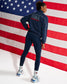 A woman stands with her back to the camera wearing a navy blue USA Wrestling track jacket and matching leggings, posed in front of a large American flag backdrop.