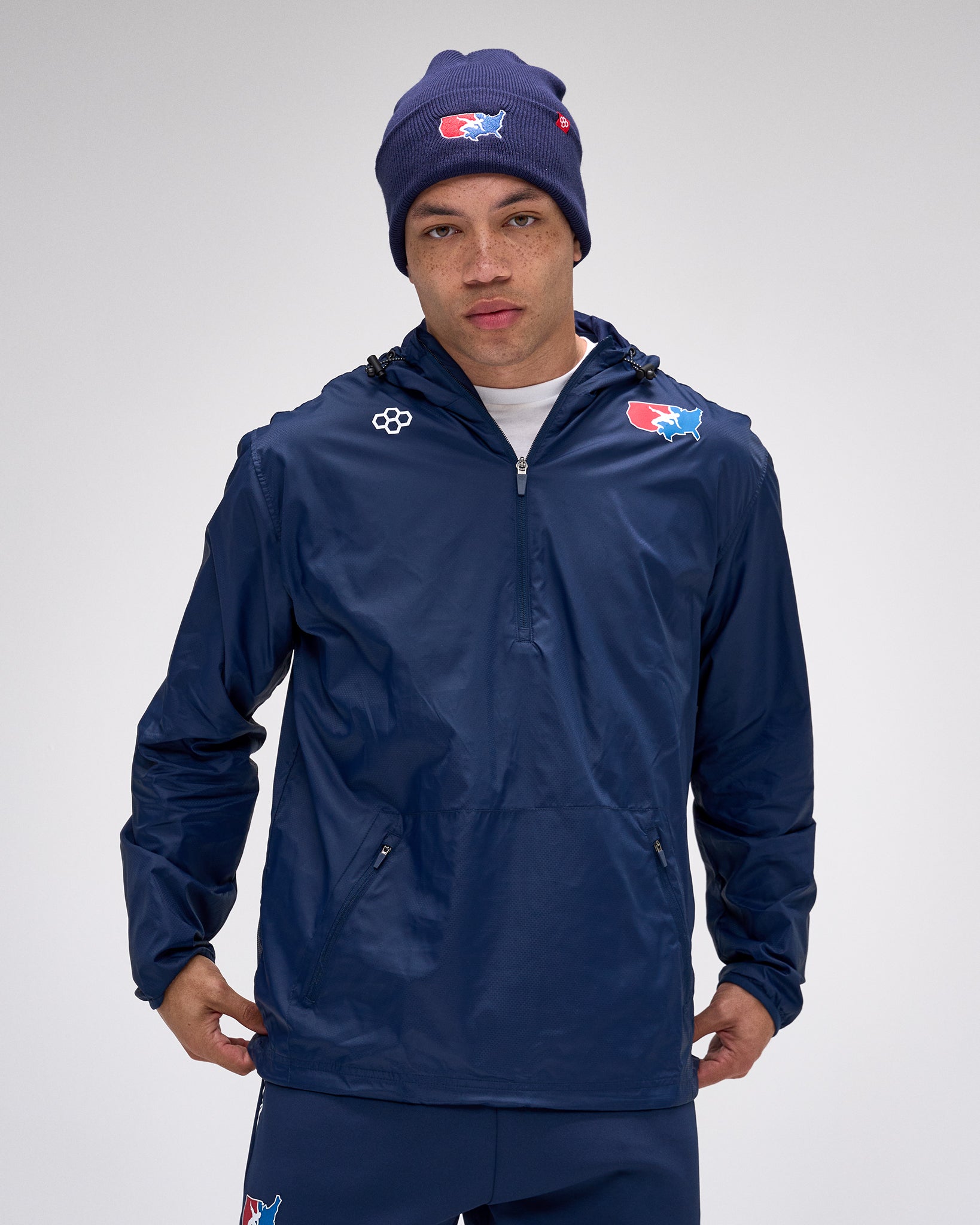 Person wearing a navy blue jacket and matching beanie with logos on a gray background