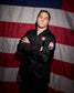 A man wearing a black jacket with patriotic red, white, and blue patches stands with arms crossed in front of a large American flag backdrop.