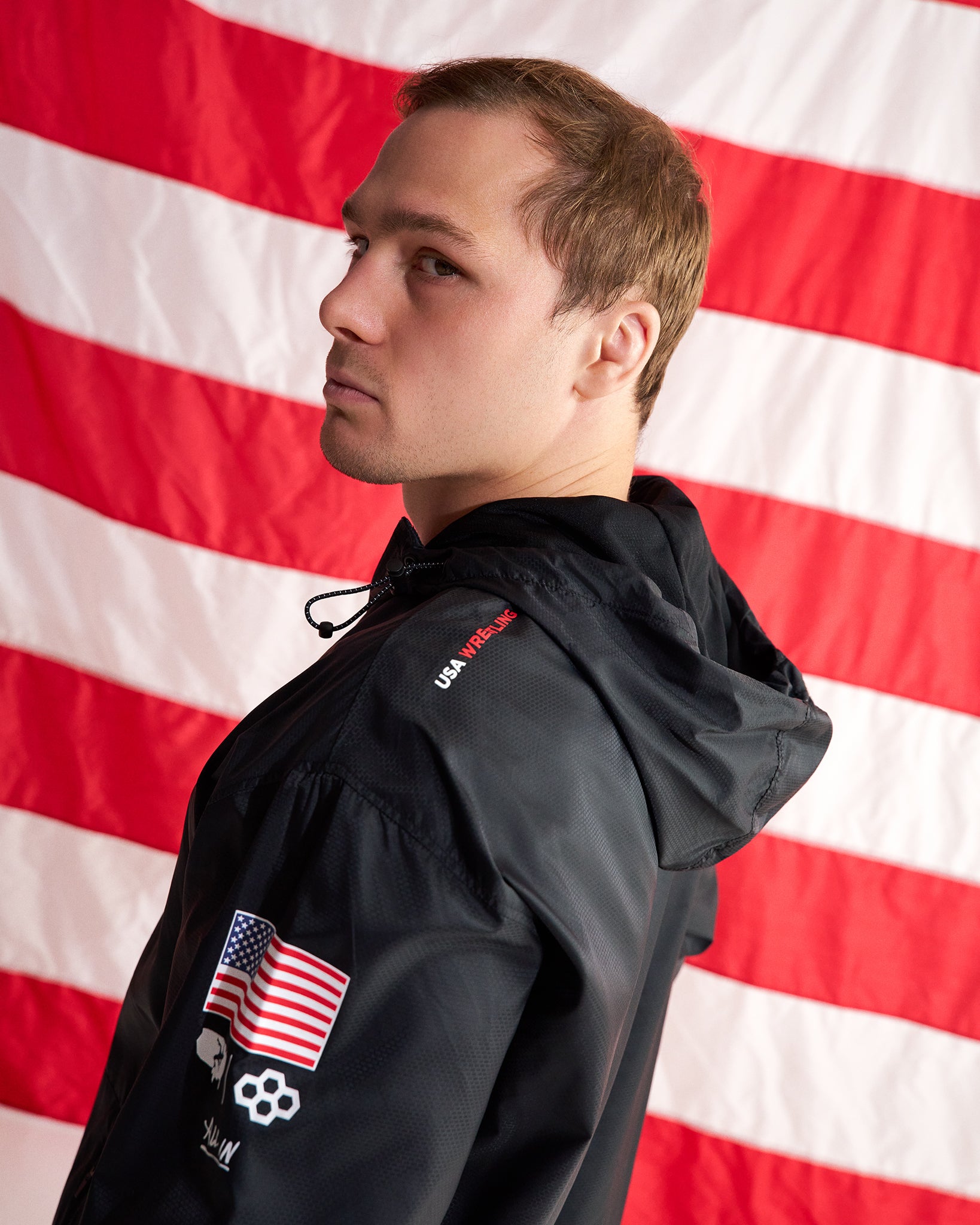 A man in profile wearing a black hooded jacket with a US flag patch and skull logo on the sleeve, posed against a large American flag backdrop.