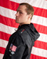 A man in profile wearing a black hooded jacket with a US flag patch and skull logo on the sleeve, posed against a large American flag backdrop.