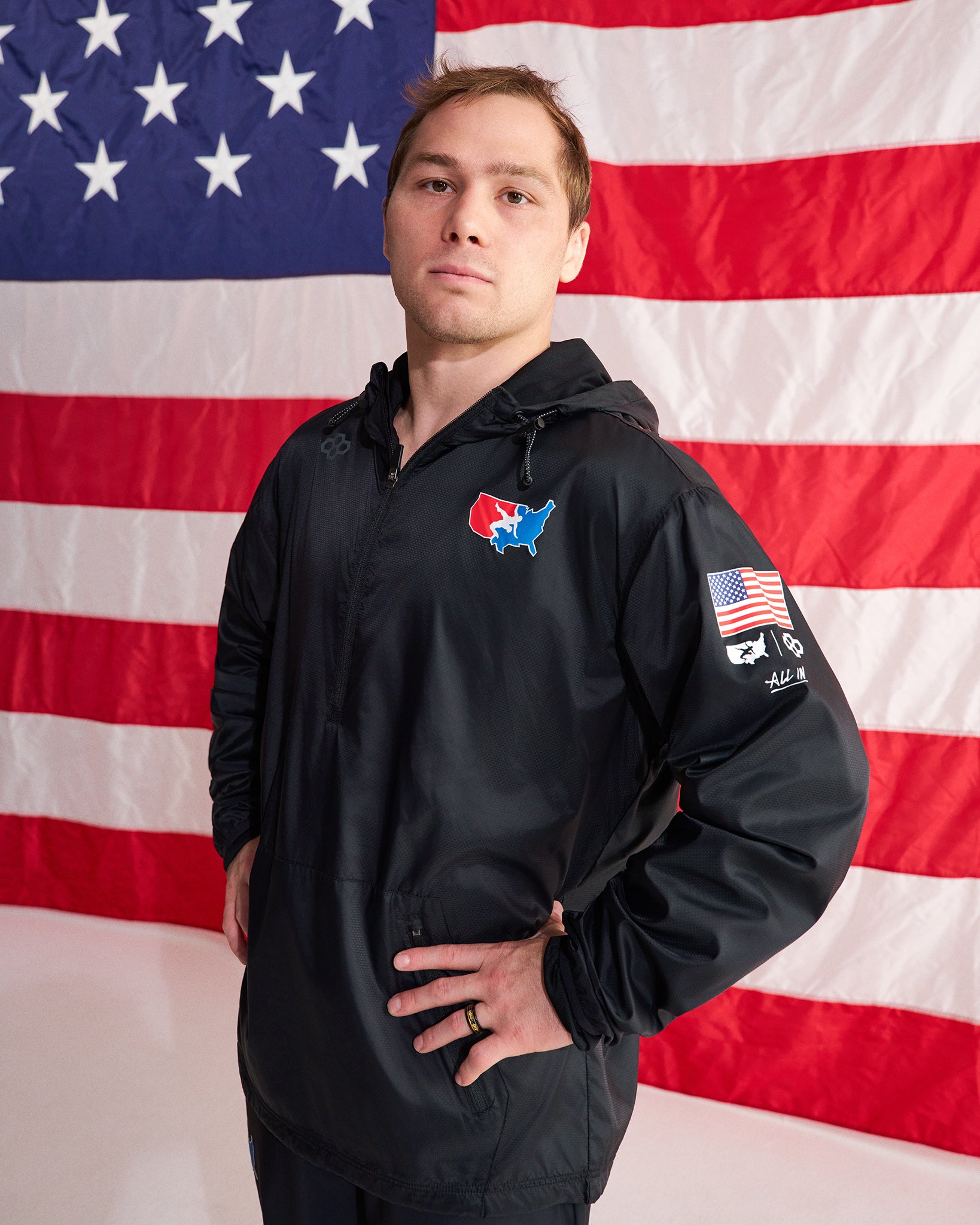 A man wearing a black hooded jacket with patriotic US-themed graphics, standing in front of a large American flag backdrop.
