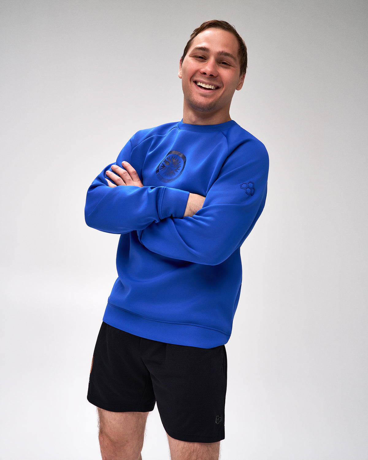 A smiling man with arms crossed wears a bright royal blue crewneck sweatshirt with a dark logo on the chest and sleeve, paired with black athletic shorts, against a light gray studio background.