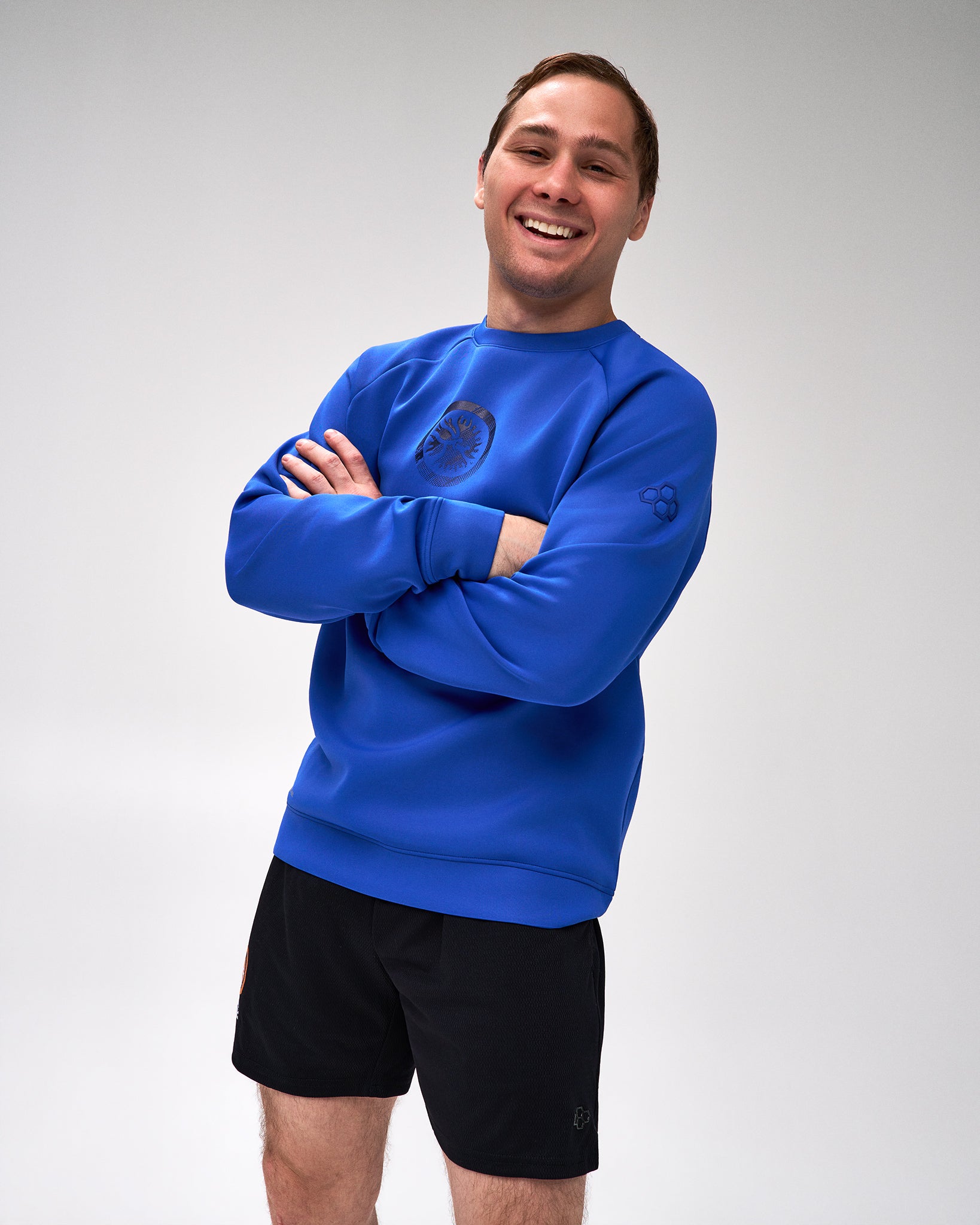 A smiling man with arms crossed wears a bright royal blue crewneck sweatshirt with a dark logo on the chest and sleeve, paired with black athletic shorts, against a light gray studio background.