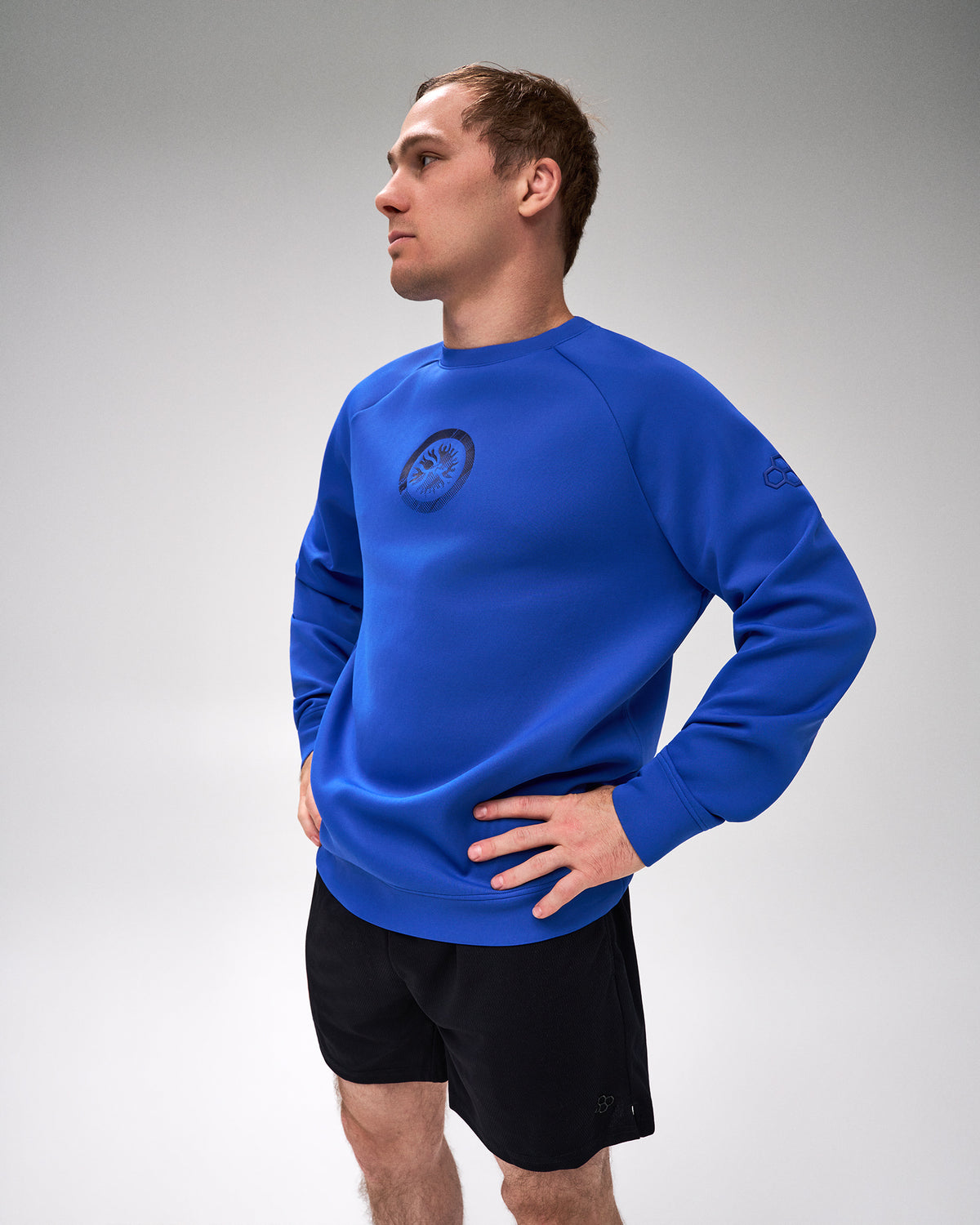 A man wearing a royal blue long-sleeve athletic crewneck sweatshirt with a circular logo on the left chest, paired with black athletic shorts, standing against a light gray studio background.