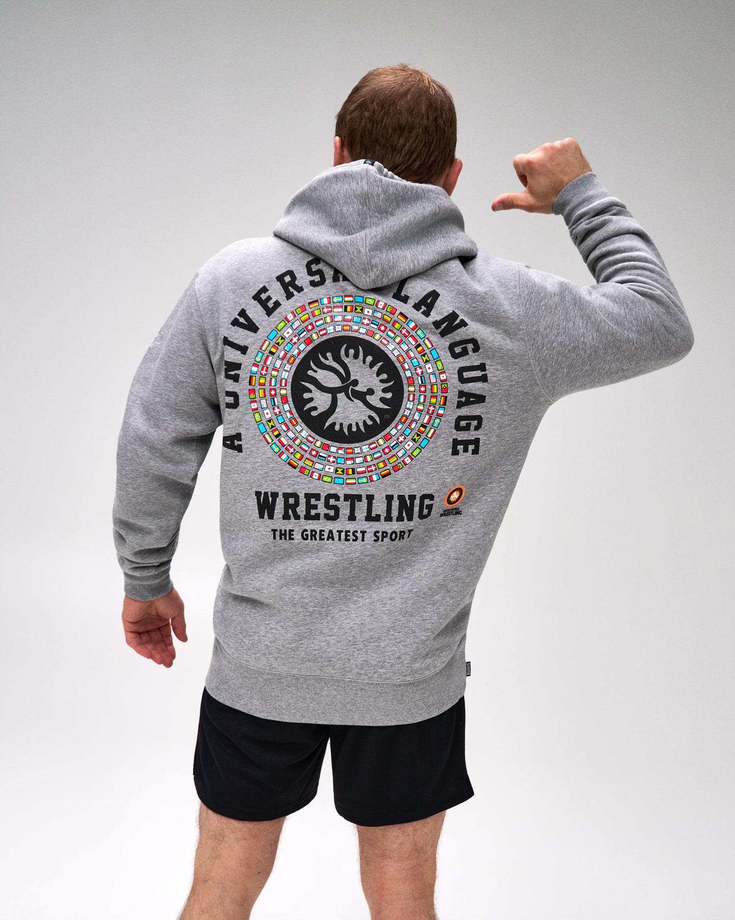 A man wearing a heather gray hoodie with a large back graphic that reads 'UNIVERSAL LANGUAGE WRESTLING THE GREATEST SPORT' and a circular multicolor emblem, shown from the back against a light gray studio background.