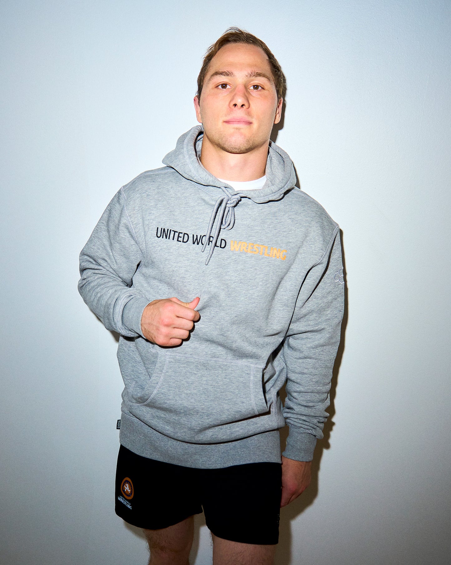 A man wearing a heather gray pullover hoodie with 'UNITED WORLD WRESTLING' printed across the chest in black and yellow, paired with black athletic shorts, standing against a plain light gray wall.