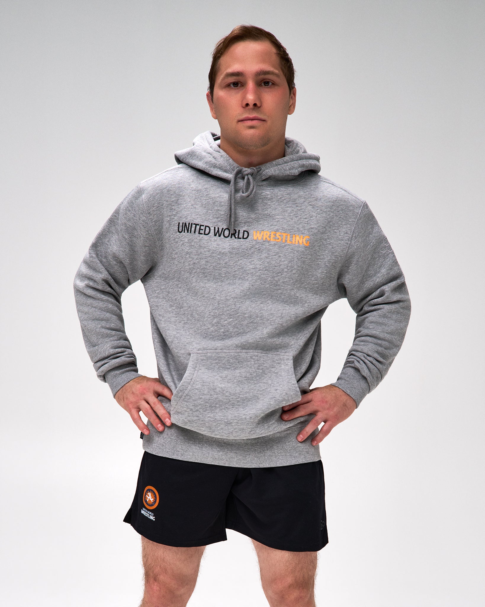 A man modeling a heather gray pullover hoodie with 'UNITED WORLD WRESTLING' printed across the chest in black and orange, paired with black athletic shorts, against a light gray studio background.