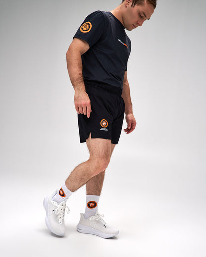 A man wearing a dark navy athletic t-shirt and matching shorts with orange and white circular logos, paired with white athletic socks and white sneakers, walking against a light gray studio background.