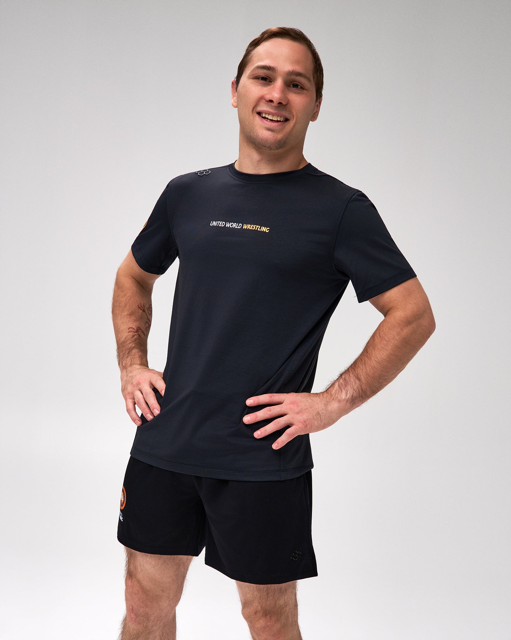 A man modeling a black short-sleeve athletic t-shirt with 'United World Wrestling' printed in gold on the chest, paired with black athletic shorts, against a light gray studio background.
