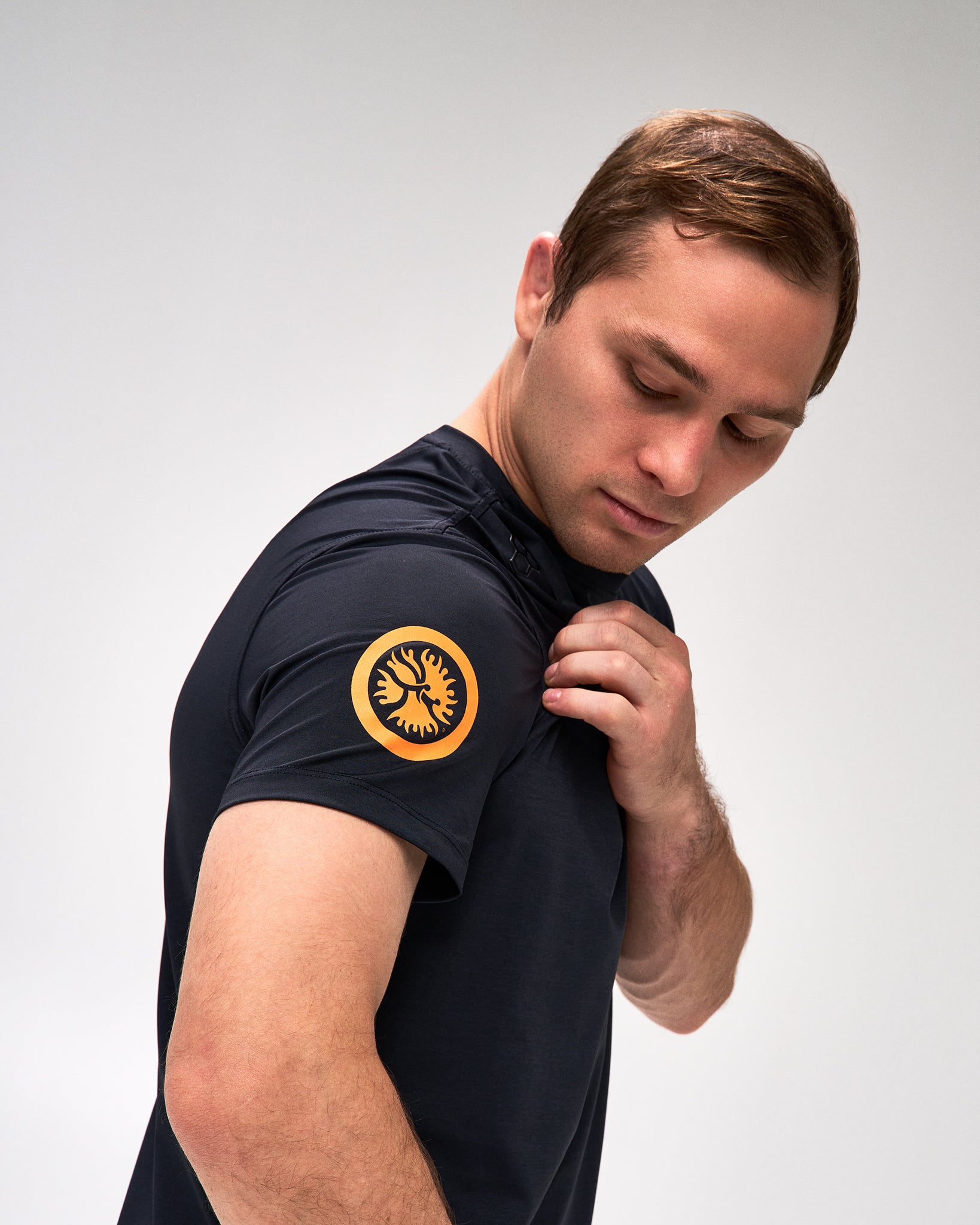 A man wearing a dark black athletic t-shirt with a circular yellow and black UWW logo patch on the left sleeve, shown in a studio setting against a light gray background.