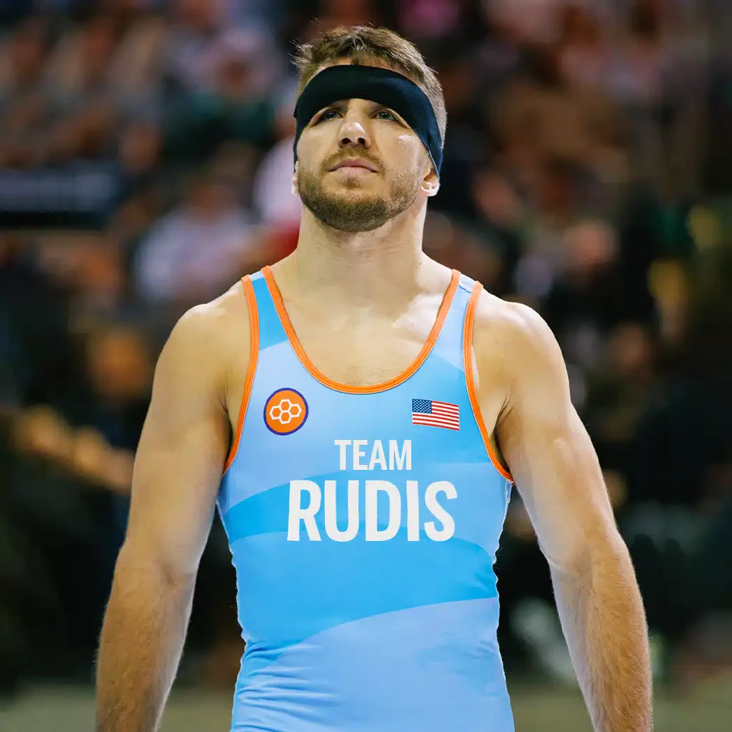 A focused male wrestler in a blue singlet representing Team Rudis wearing a black headband and displaying determination before a match