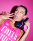 Athlete wearing a pink RUDIS singlet poses against a vibrant pink backdrop while holding a black and pink RUDIS wrestling shoe, showcasing bold style, matching pink nail art, and expressive personality.
