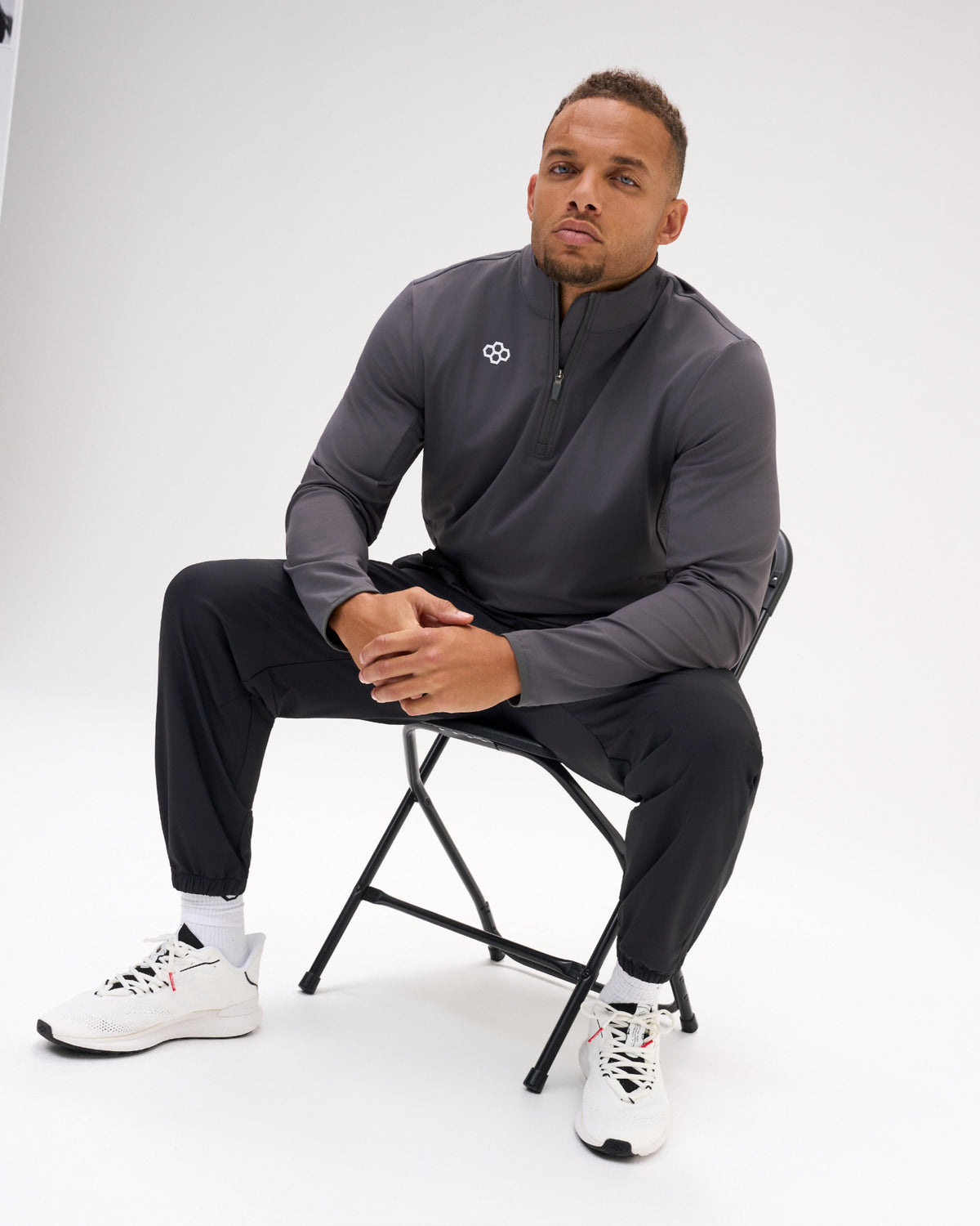Man wearing the carbon RUDIS Victory 1/4 Zip with black athletic pants and white training shoes in a seated studio setting.